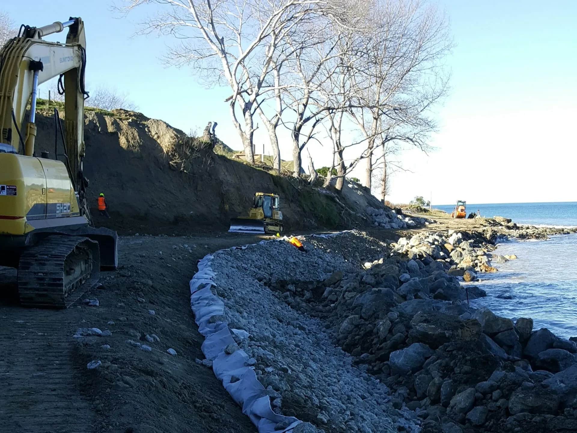 Excavator and bulldozer placing rock armour along the eroding coastal bank at Anaura Bay, Tairāwhiti