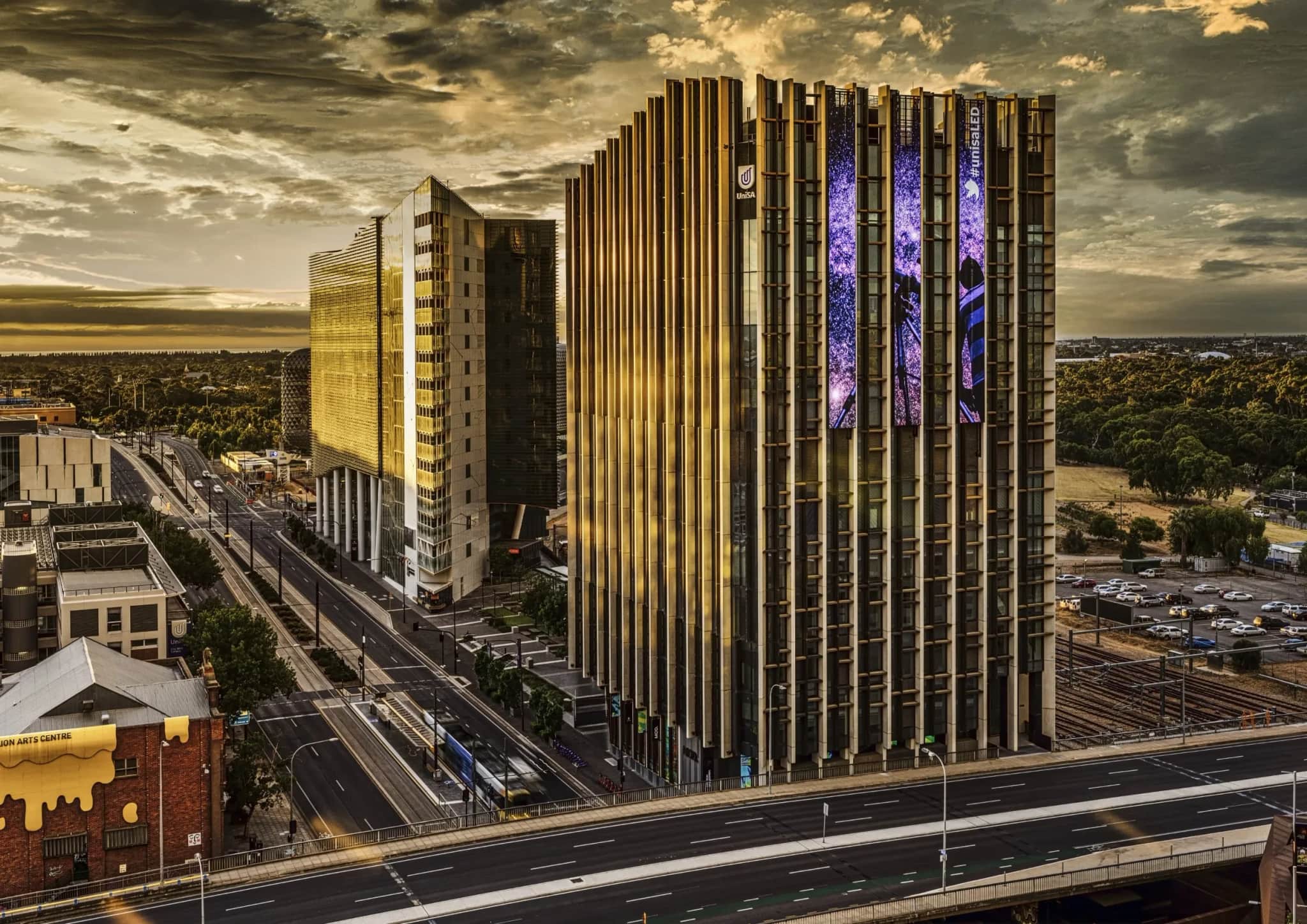 The University of South Australia's Bradley Building at dusk in Adelaide's West End, with two tall vertical LED screens lit on its façade above the tramline.
