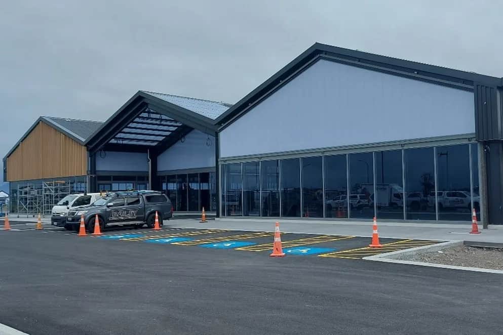 Nearly completed building frontage with timber-clad and grey gabled sections, a glazed canopy entrance and full-height glazing.