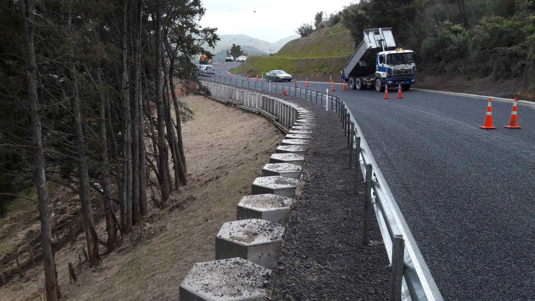 A new concrete retaining wall and guardrail on a rural road, with a tipper truck and road cones