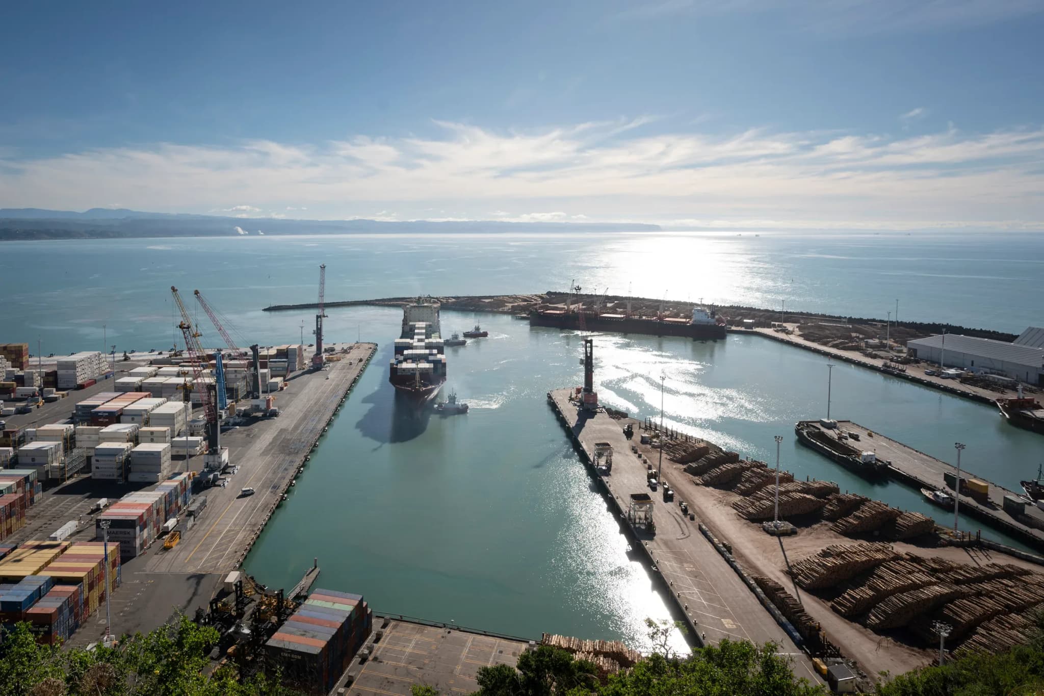 Container ship under tow between the wharves at Napier Port, with log stacks, containers and Hawke Bay behind