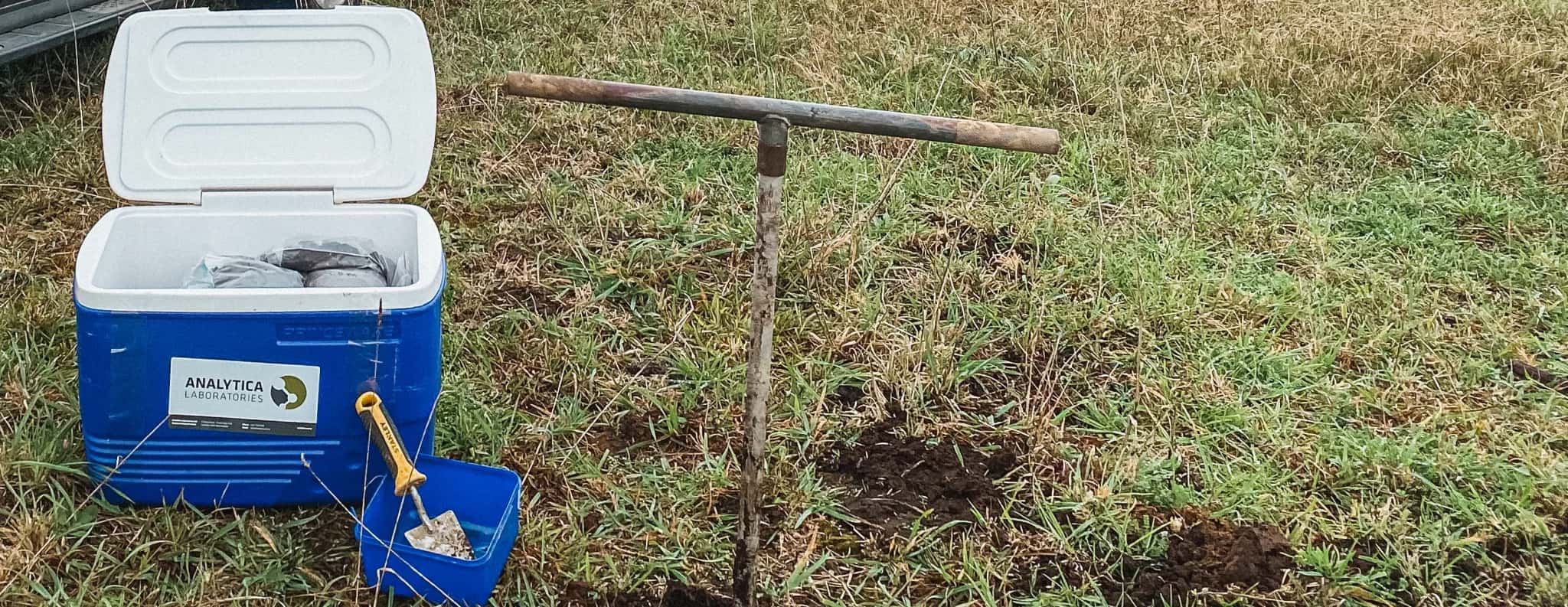 A hand auger in a soil sampling hole beside a laboratory sample chilly bin